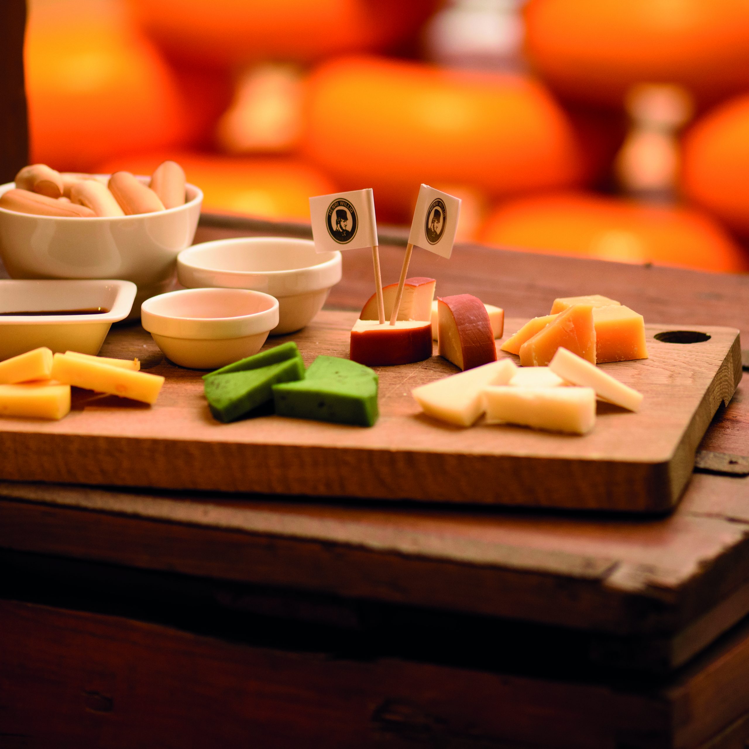 Cheese tasting: A wooden plank featuring four slices of Dutch cheese. The cheeses vary in color, texture, and shape, inviting viewers to appreciate their diversity. Each slice is accompanied by a small knife for serving. The aroma emanating from the cheeses hints at their rich flavor profiles, enticing onlookers to enjoy a tasting experience.