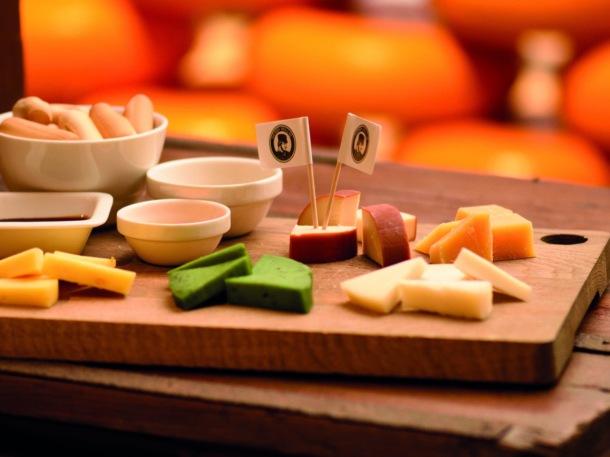 Cheese tasting: A wooden plank featuring four slices of Dutch cheese. The cheeses vary in color, texture, and shape, inviting viewers to appreciate their diversity. Each slice is accompanied by a small knife for serving. The aroma emanating from the cheeses hints at their rich flavor profiles, enticing onlookers to enjoy a tasting experience. Comprehensive Guide to the Bollenstreek